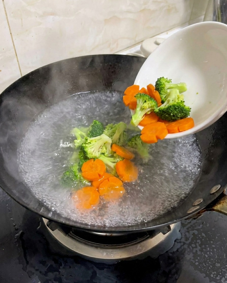 Brócoli fresco y zanahorias en forma de flor vertiéndose desde un tazón blanco a un wok con agua hirviendo.