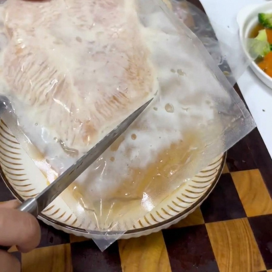 Una mano usando un cuchillo para abrir un paquete de plástico transparente al vacío que contiene una pieza de carne de pollo marinada.