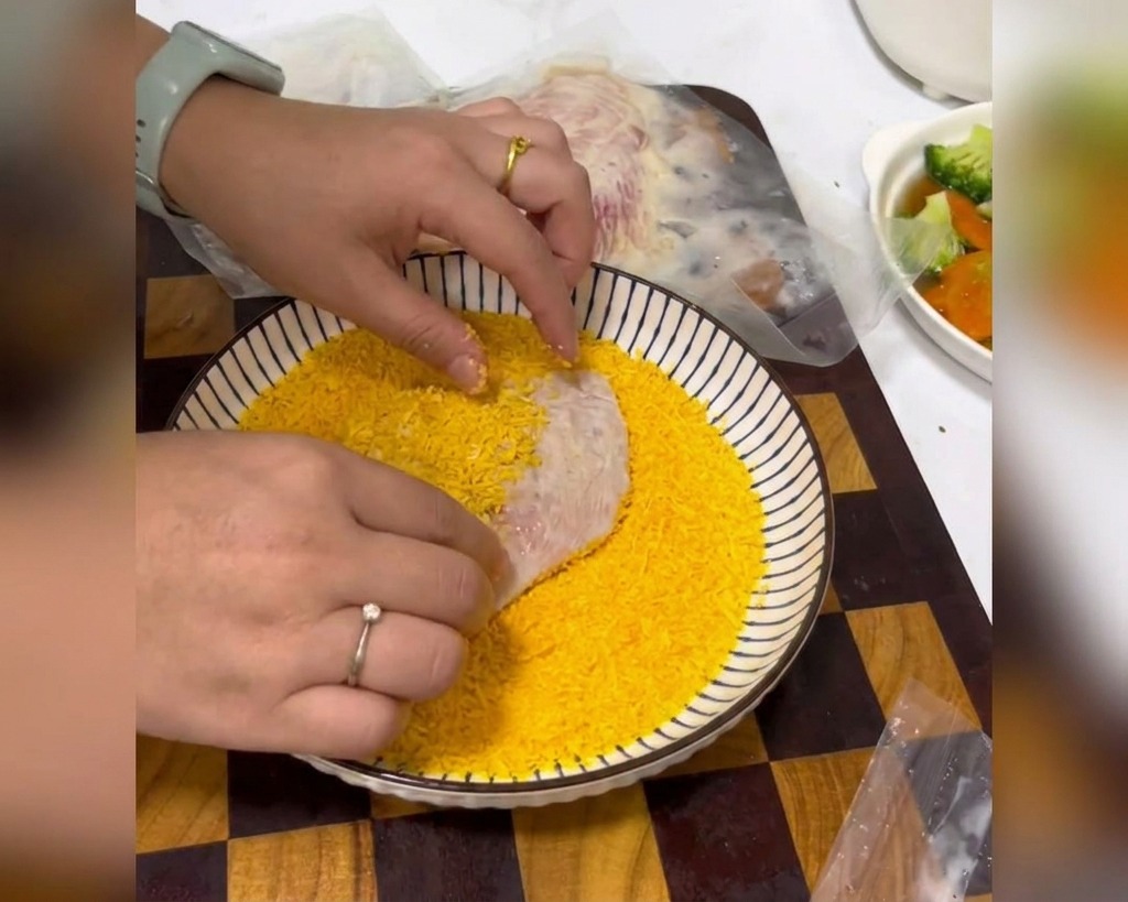 Manos presionando una pechuga de pollo marinada en un tazón blanco poco profundo lleno de pan rallado amarillo brillante.