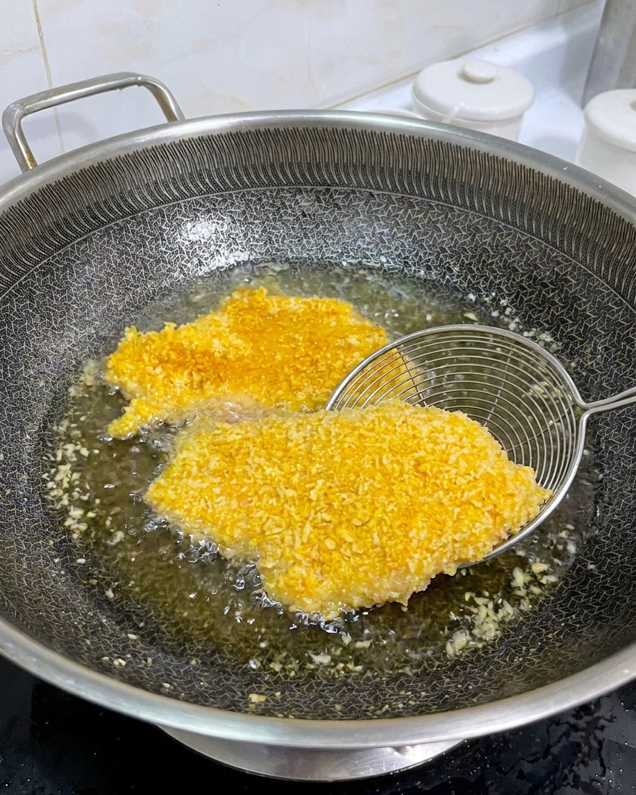 Un colador de metal levantando una chuleta de pollo empanizada y dorada en una sartén con aceite caliente para mostrar el lado cocinado.