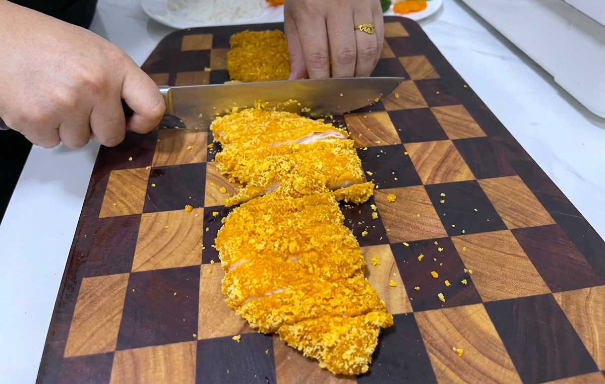 Una persona usando un cuchillo grande para cortar un filete de pollo empanizado, crujiente y dorado sobre una tabla de cortar de madera oscura.