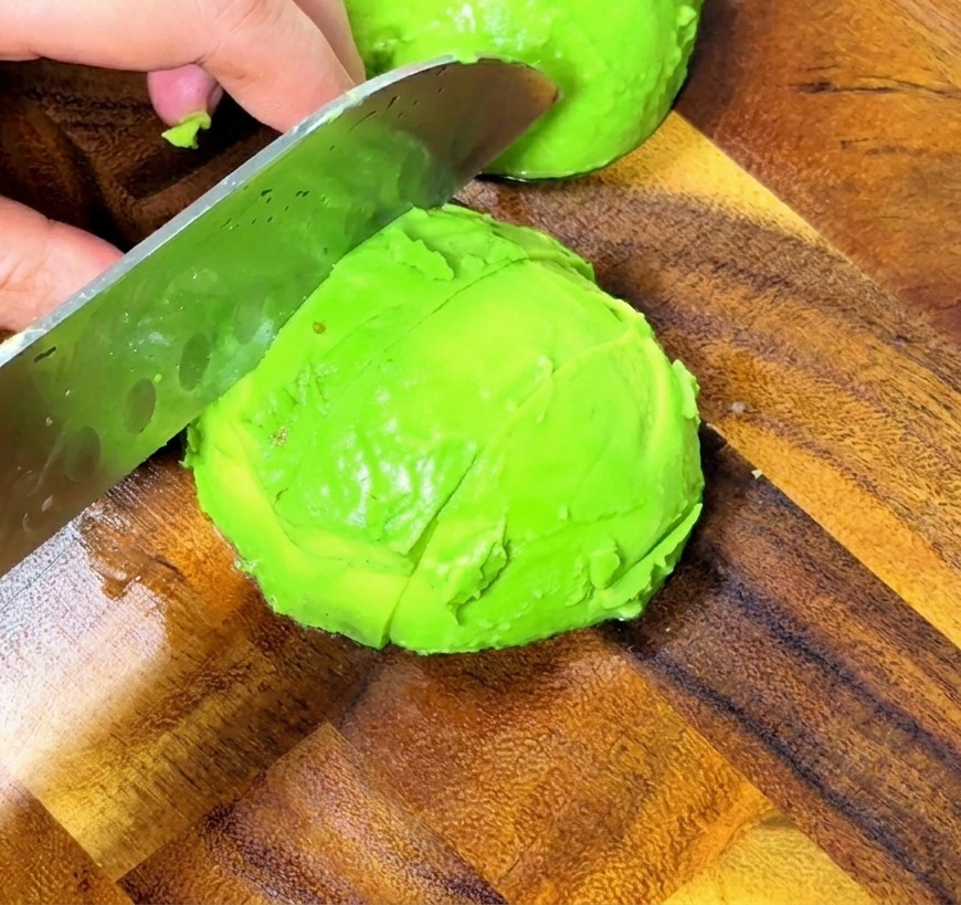 Primer plano de unas manos cortando un aguacate pelado en cubos uniformes sobre una tabla de madera usando un cuchillo profesional.