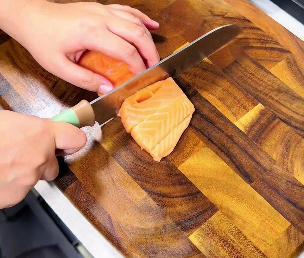 Manos cortando un filete de salmón fresco sobre una tabla de madera para crear cubos uniformes del tamaño de un bocado.