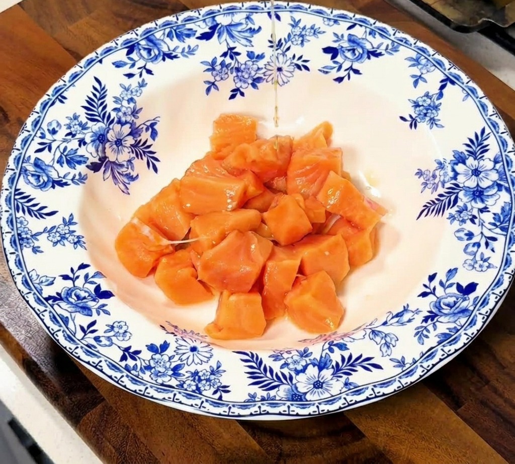 Líquido oscuro de marinada vertiéndose sobre un montoncito de dados de salmón crudo en un tazón de cerámica con motivos florales.