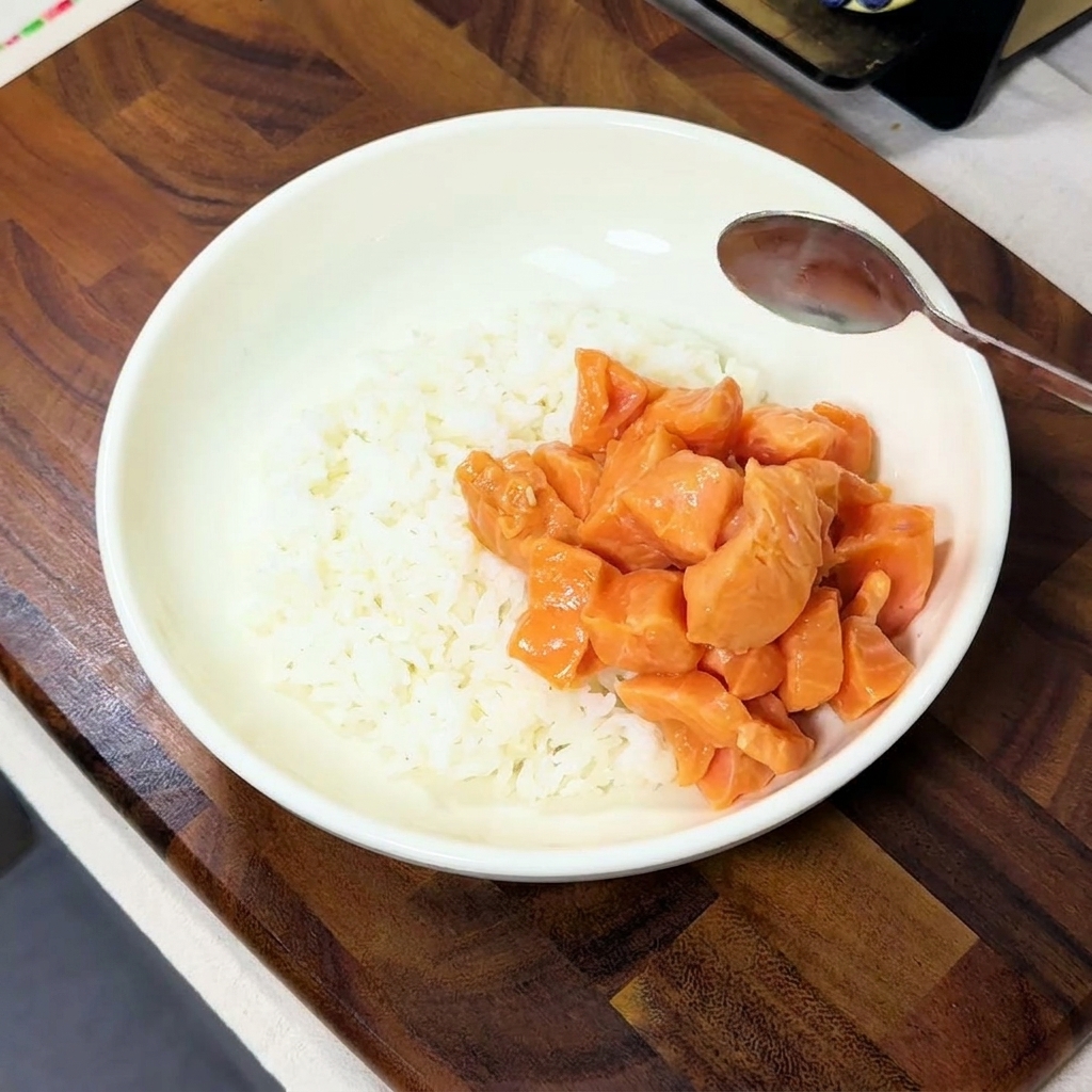 Dados de salmón marinado colocándose con una cuchara en un lado de un tazón blanco lleno de arroz cocido al vapor.
