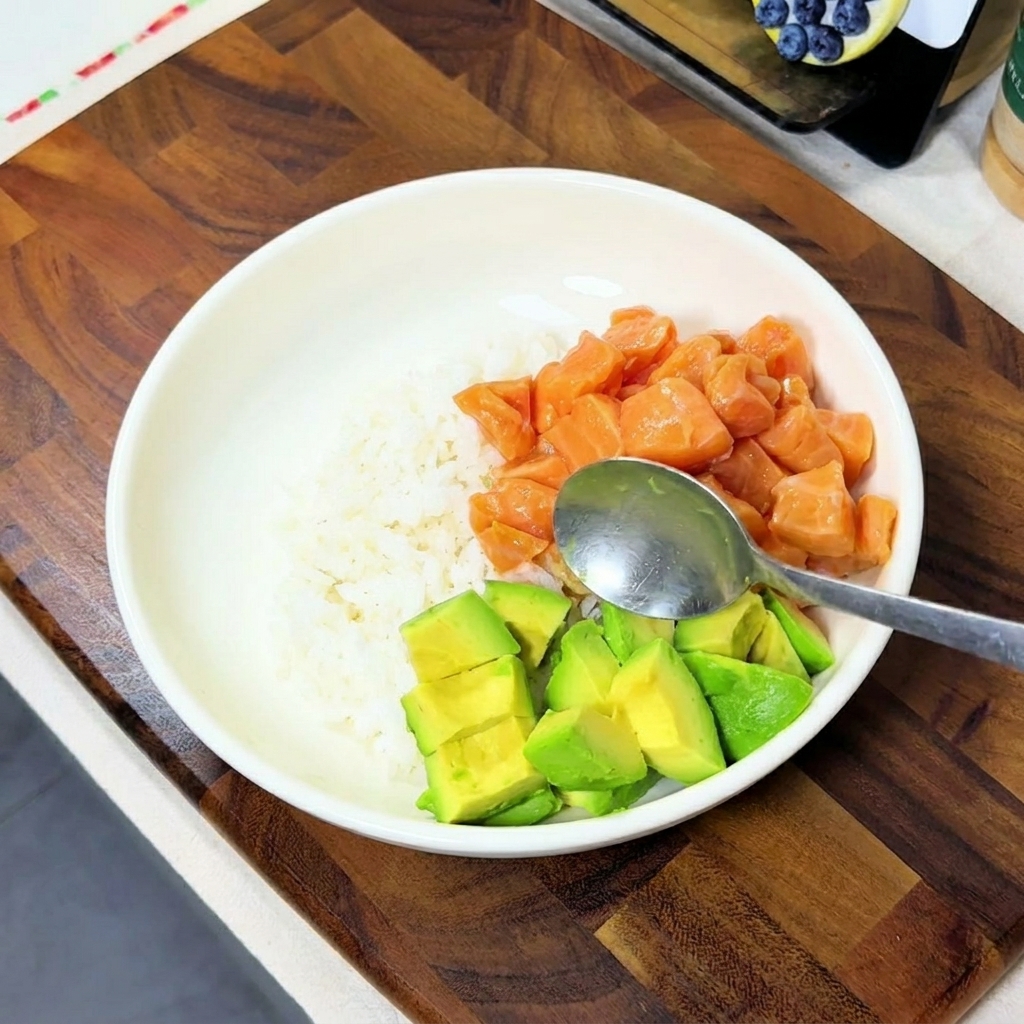 Dados de aguacate añadiéndose con una cuchara a un tazón blanco que ya contiene arroz y salmón marinado.