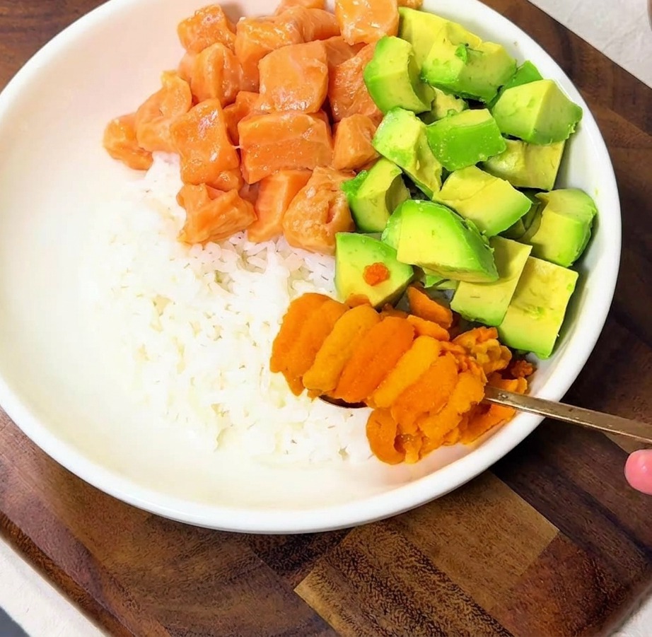 Una mano colocando trozos de erizo de mar de un naranja brillante sobre un tazón de arroz que ya tiene salmón y aguacate.