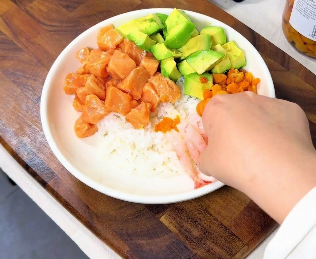 Una mano acomodando varios camarones dulces rosados y translúcidos a un lado del tazón de arroz.