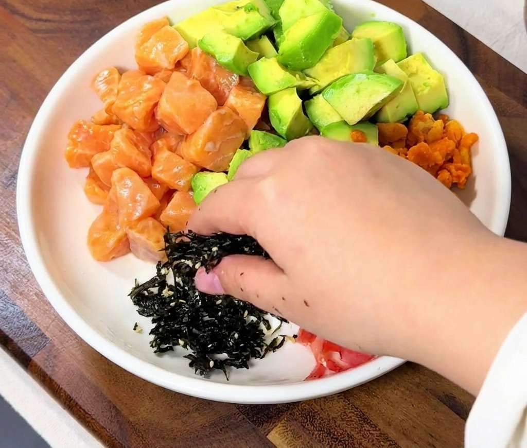 Una mano espolvoreando un montoncito de algas negras en tiras sobre el tazón de arroz junto a los dados de salmón.