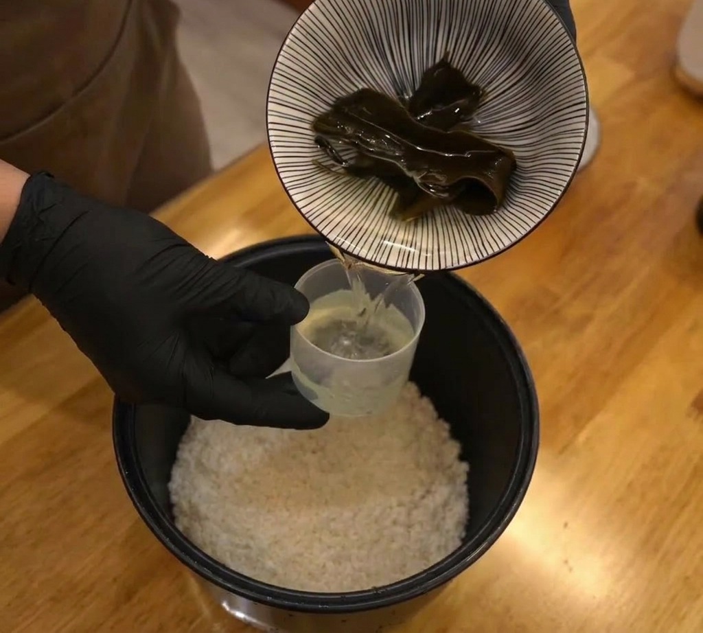 Un trozo oscuro de alga kombu seca colocándose en una arrocera llena de arroz blanco y agua.