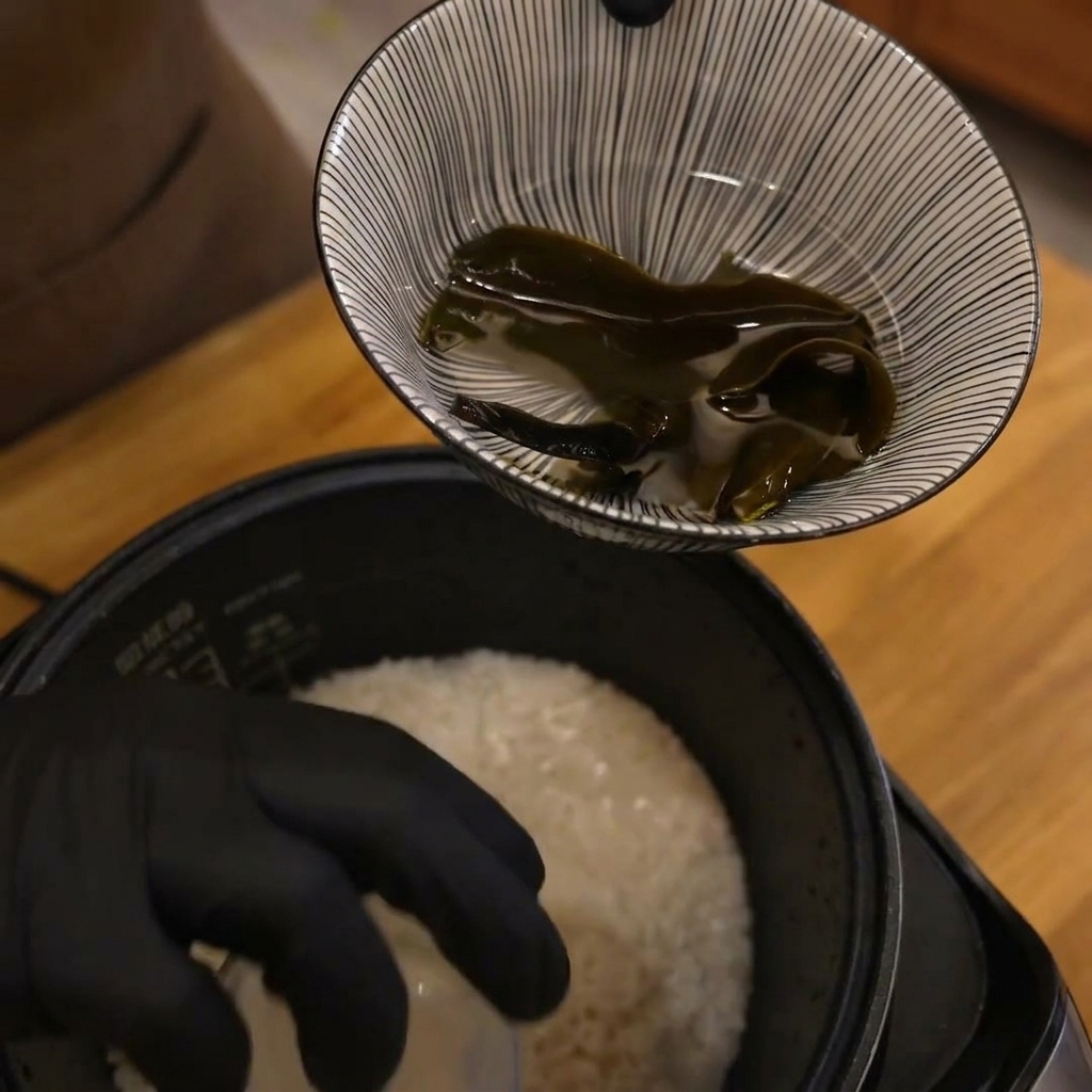 Una mano con guante sosteniendo una taza medidora sobre una olla de arroz blanco antes de añadir agua.
