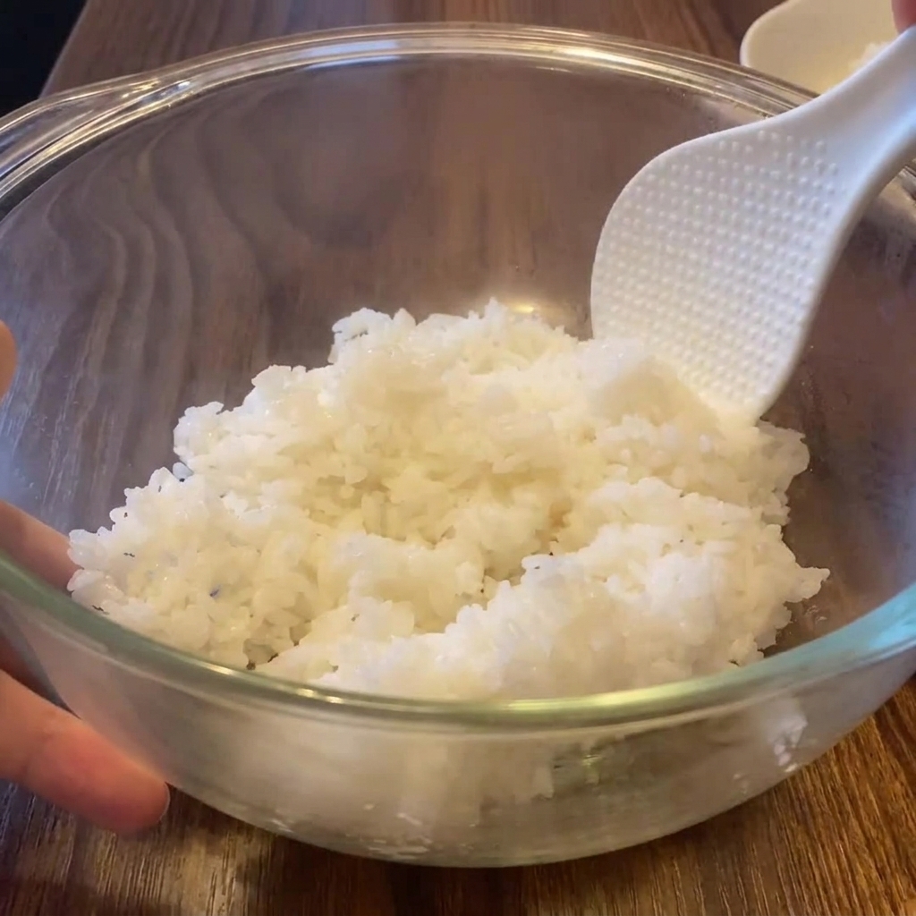 Una espátula de arroz ahuecando arroz blanco humeante dentro de un bol de cristal grande.