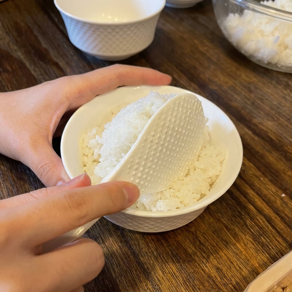 Una espátula de arroz colocando una porción de arroz blanco en un tazón de cerámica blanca.