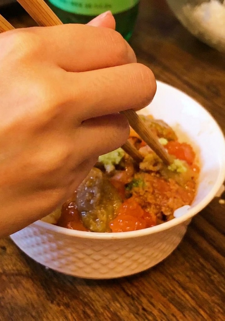 Una mano usando palillos para colocar una pequeña cantidad de wasabi en el centro de un tazón de arroz con erizo y salmón.