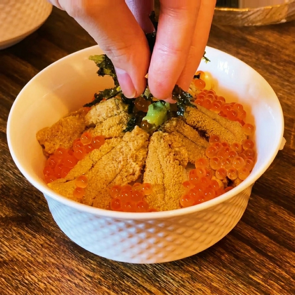Dedos espolvoreando alga nori oscura en tiras sobre la parte superior de un tazón de sushi con erizo y huevas.