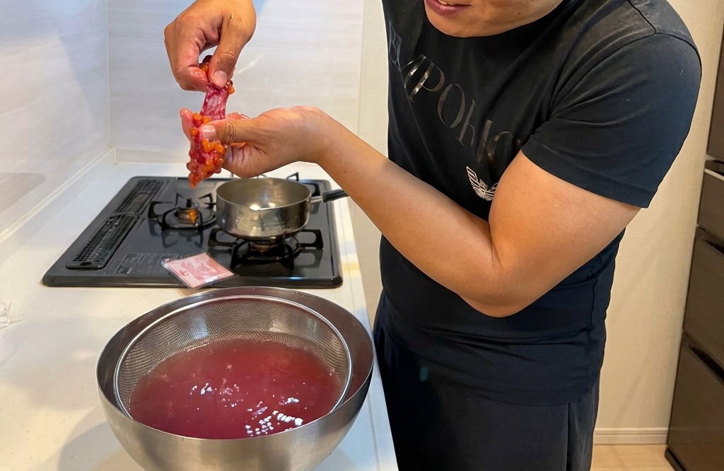 Manos frotando suavemente el saco de huevas contra un tamiz metálico sumergido en agua tibia.