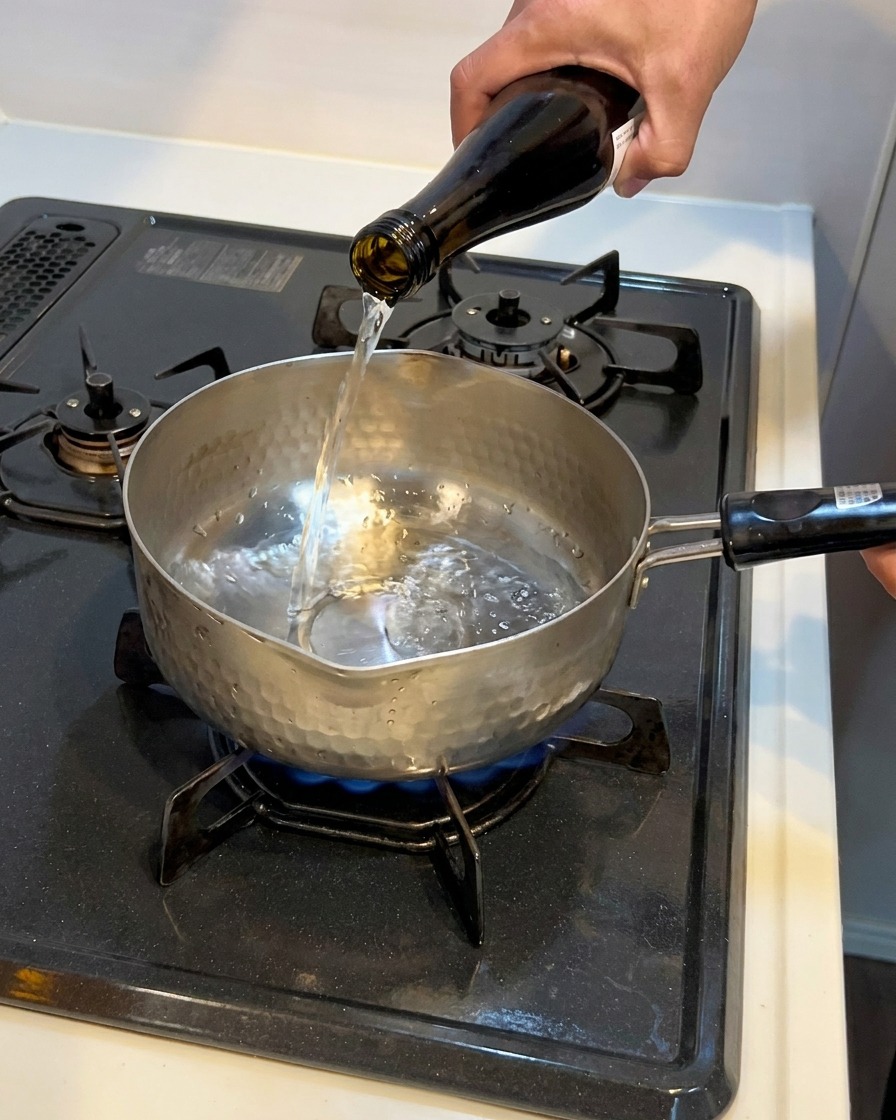Líquido transparente vertiéndose desde una botella de vidrio oscuro en una pequeña olla de metal.