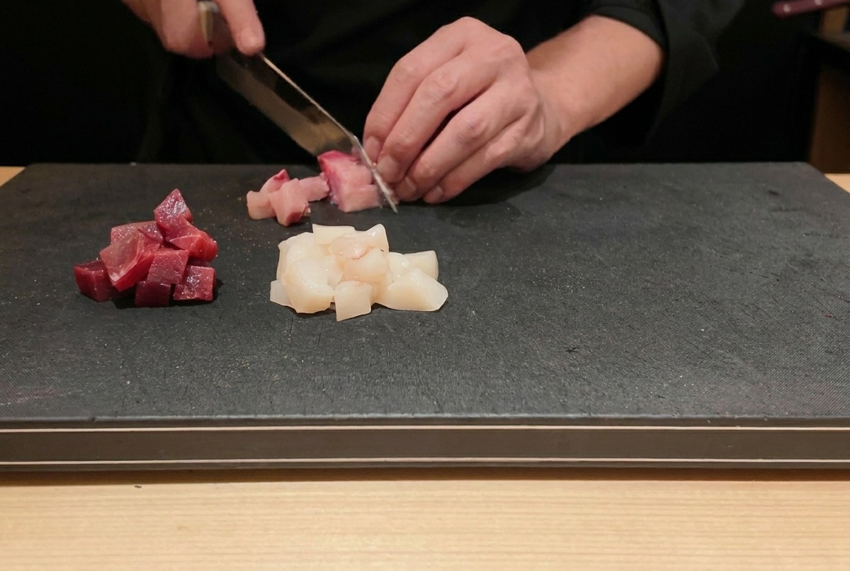 Un chef cortando atún fresco crudo y pez limón en cubos pequeños sobre una tabla de cortar negra.