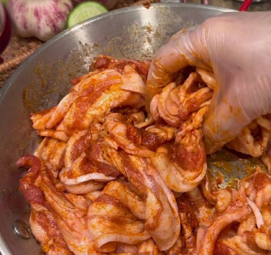 Una mano con guante mezclando rodajas de panceta de cerdo cruda con una marinada espesa y de color rojo oscuro en un bol de acero inoxidable.