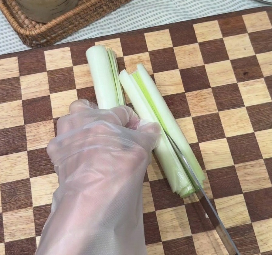 Una mano utilizando un cuchillo de chef para cortar un tallo de cebolleta blanca en tiras finas y alargadas sobre una tabla de madera a cuadros.
