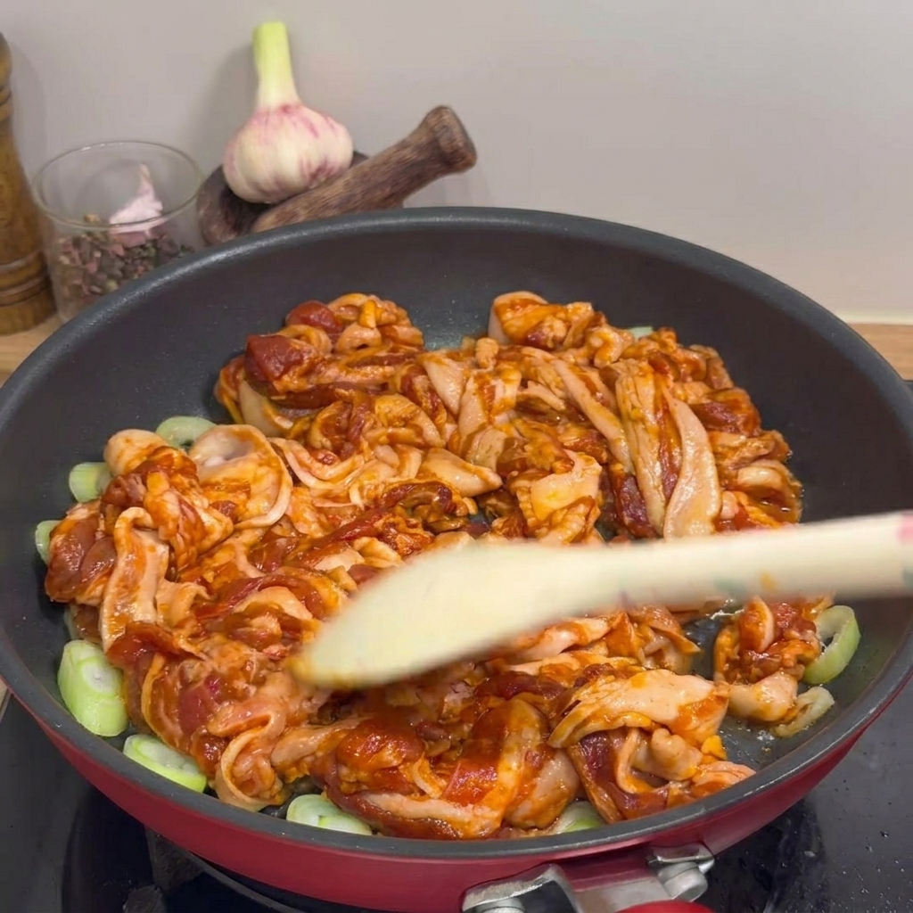 Un primer plano de tiras de panceta de cerdo marinada salteándose en una sartén roja con aros de cebolleta, usando una espátula de madera para darle la vuelta a la carne.