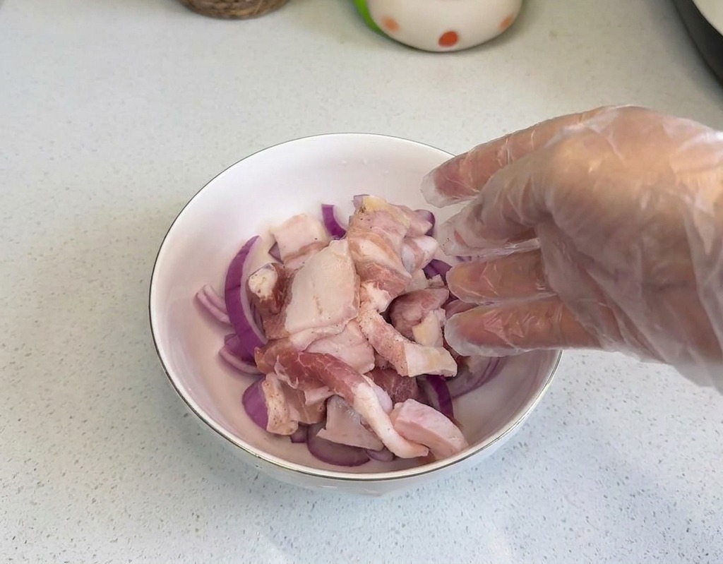 Una mano con guante de plástico colocando láminas de panceta cruda en un cuenco con aros de cebolla.