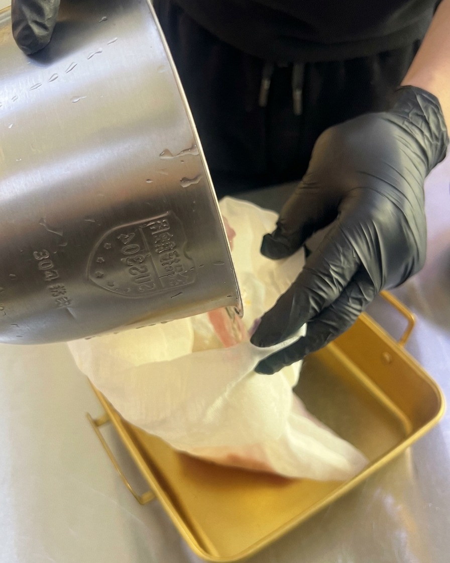 Manos con guantes exprimiendo una bolsa de tela blanca para dejar caer zumo de fruta fresco en una bandeja metálica.