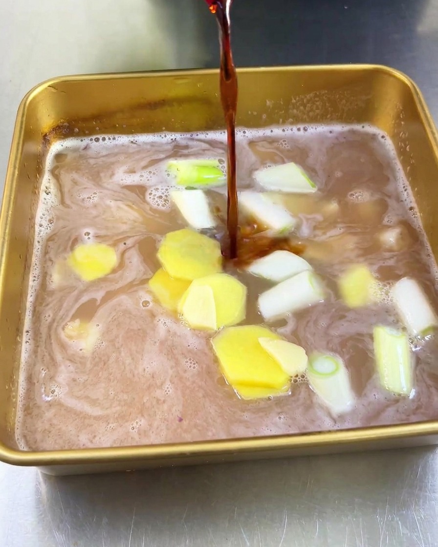 Un chorro de salsa de soja oscura vertiéndose en una bandeja con zumo de fruta claro, rodajas de jengibre y cebolleta picada.