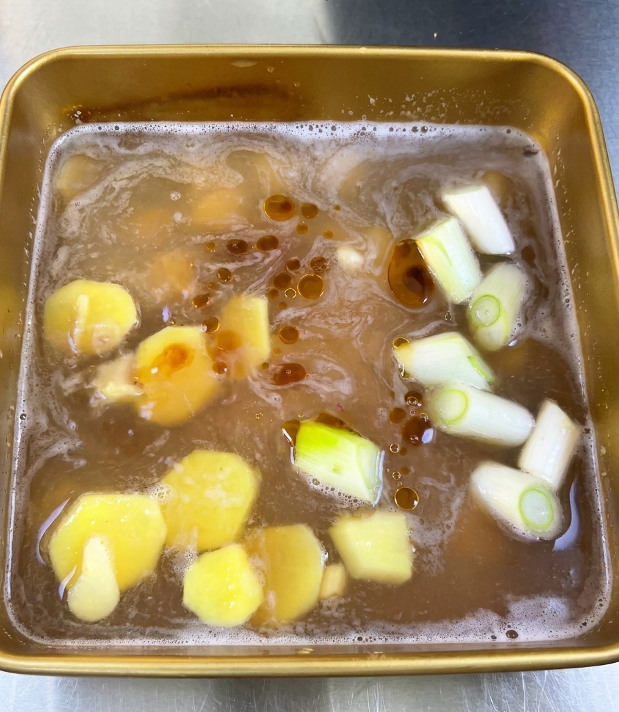 Vista cenital de un adobo de soja en una bandeja con rodajas de jengibre, cebolleta y gotas oscuras de aceite de sésamo flotando.