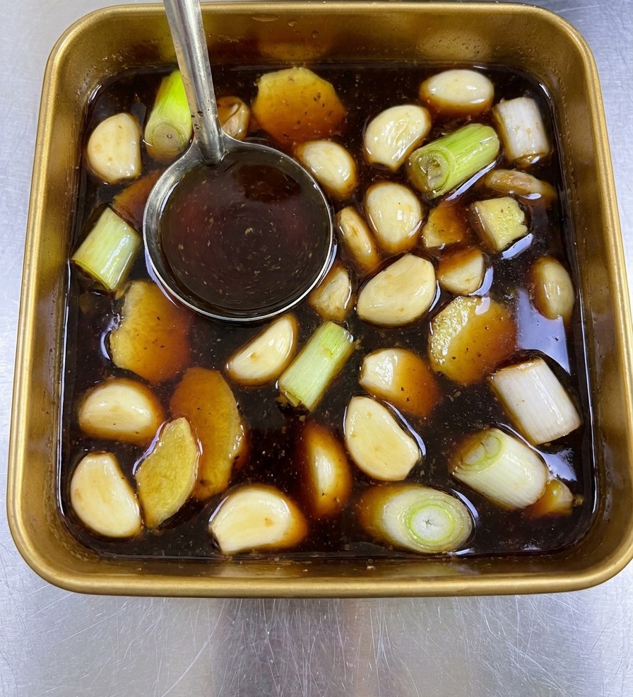 Un cucharón de metal removiendo un adobo marrón oscuro lleno de jengibre y cebolleta en una bandeja cuadrada.