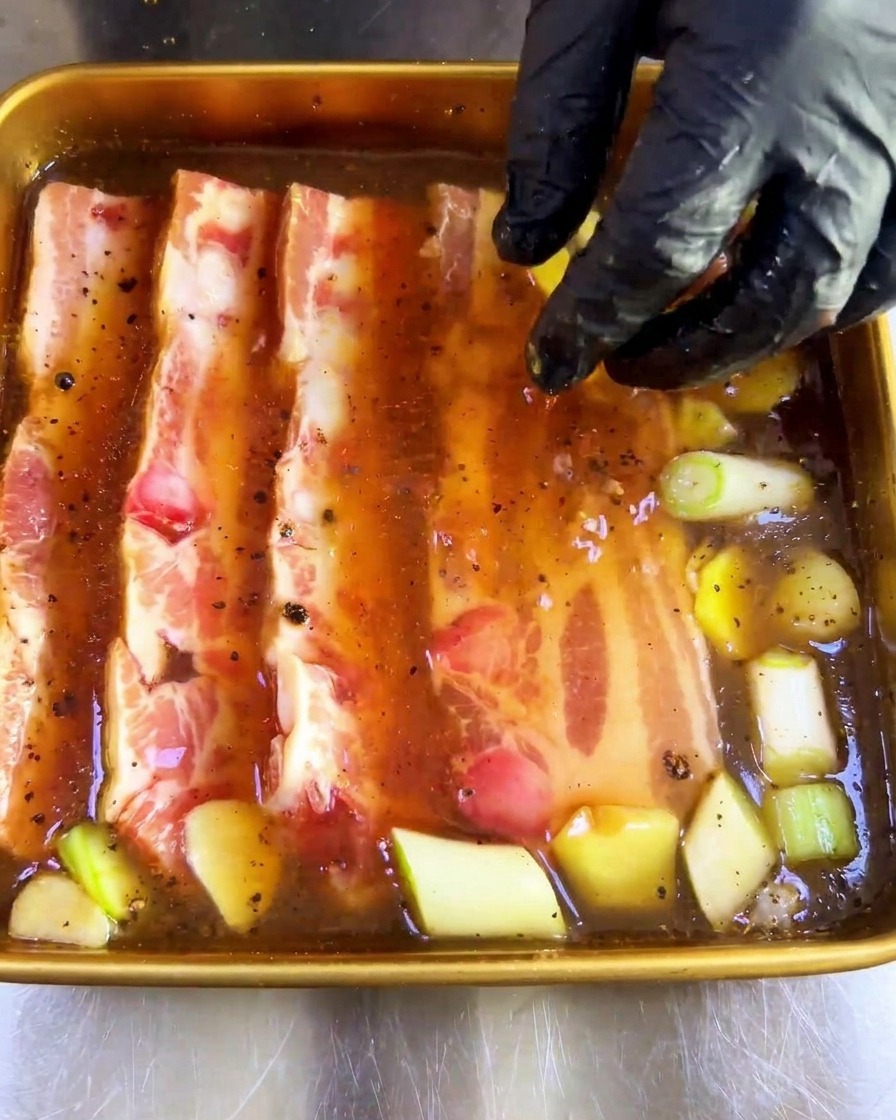 Una mano con guante presionando tiras gruesas de panceta cruda en una bandeja de adobo oscuro.
