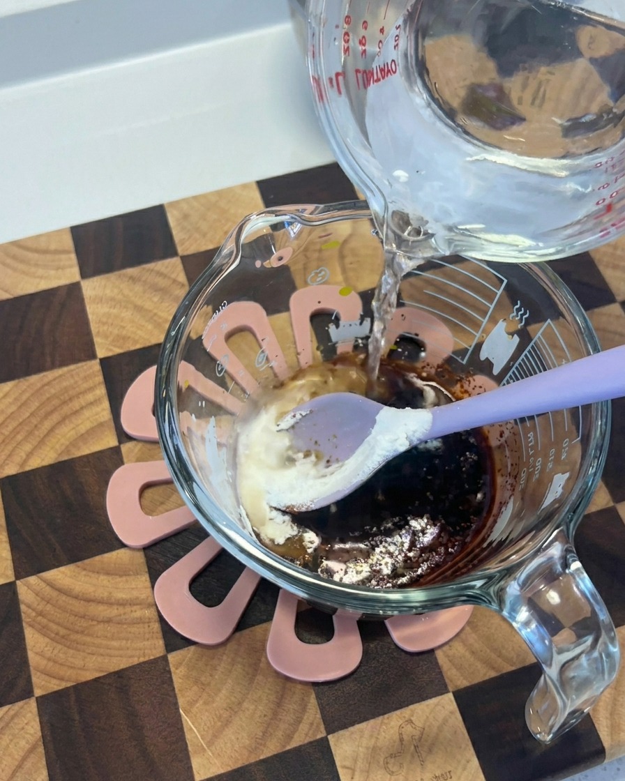 Agua vertiéndose desde un recipiente de vidrio a una taza con una mezcla oscura de salsa de soja y especias.