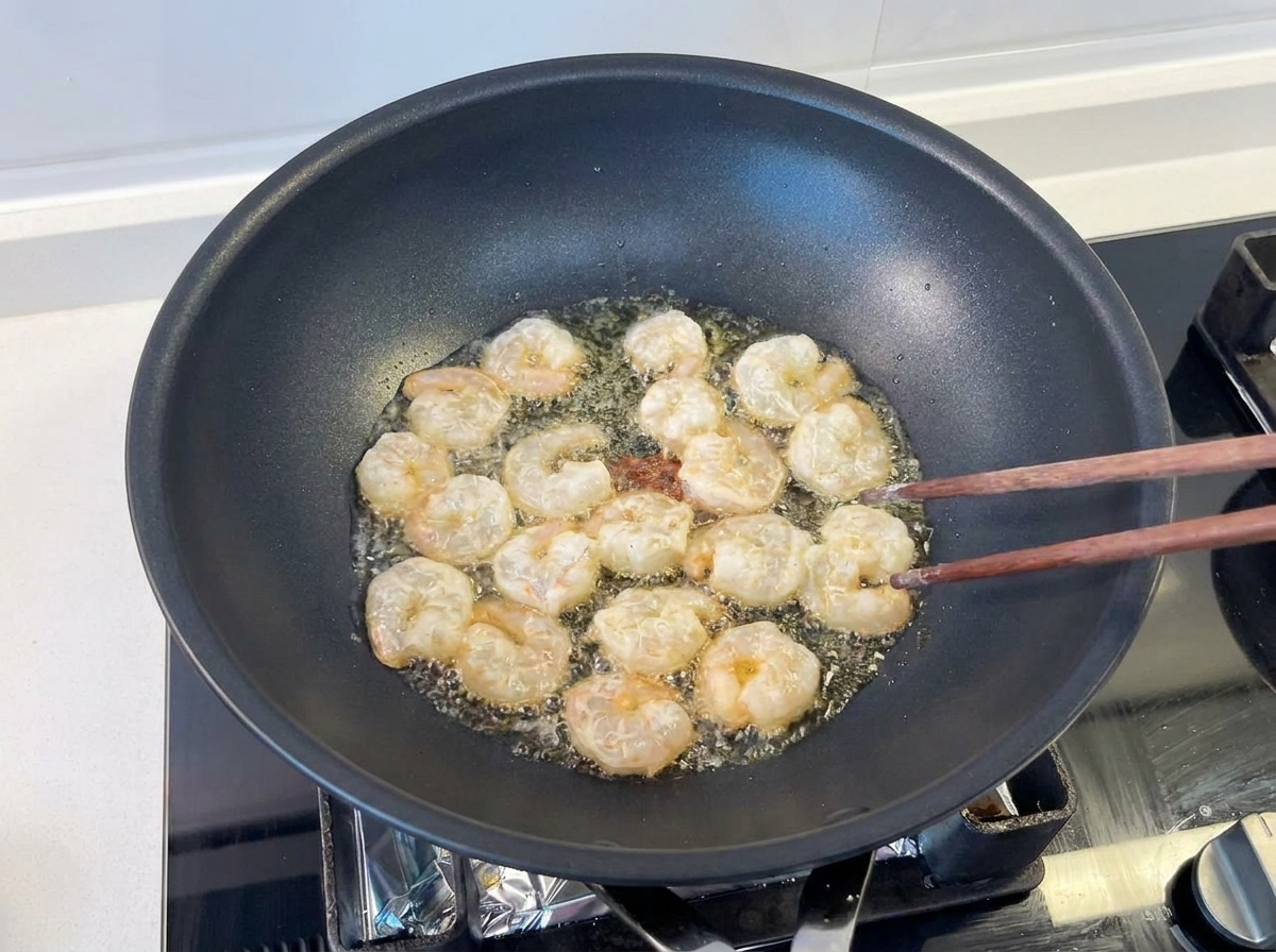 Varios camarones salteándose con palillos de madera en un wok negro con aceite brillante.