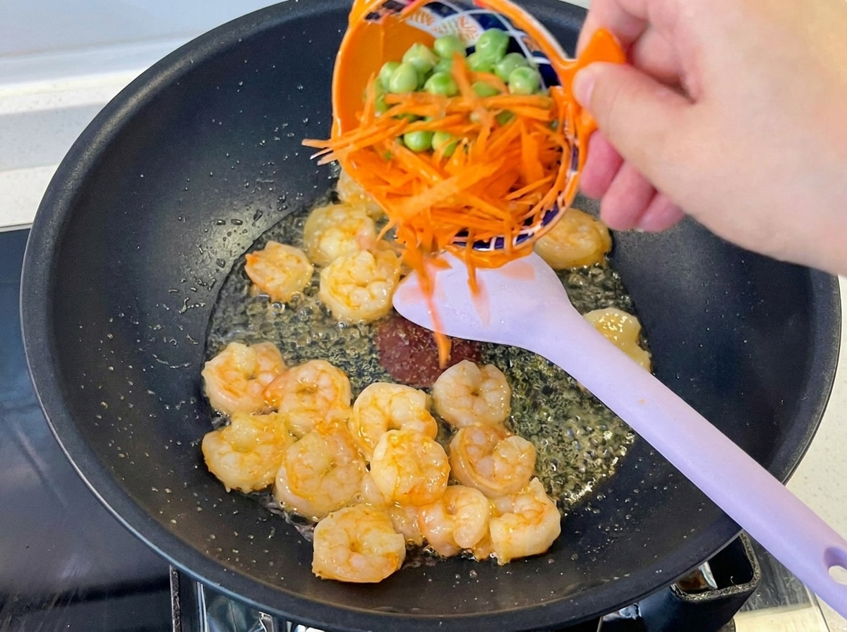 Una mano vertiendo zanahoria rallada y guisantes verdes desde un bol naranja a un wok con camarones cocidos.