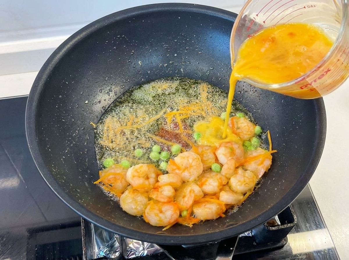 Huevos batidos vertiéndose desde una jarra de vidrio a una sartén caliente con camarones, guisantes y zanahoria.