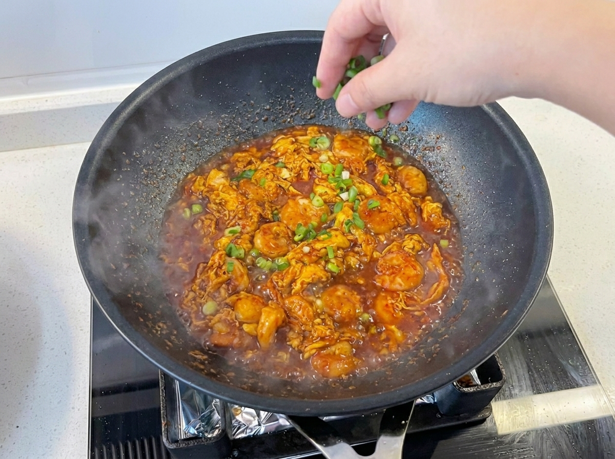Una mano esparciendo cebolleta picada fresca sobre el plato de camarones y huevos en una sartén negra.
