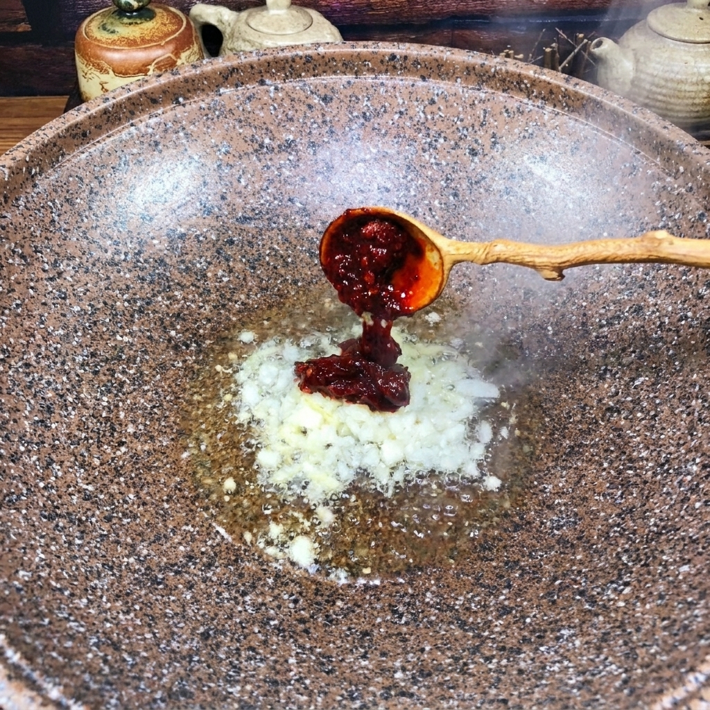 Una cuchara de madera añadiendo una porción de pasta de chile roja espesa en un wok con aromáticos salteados.
