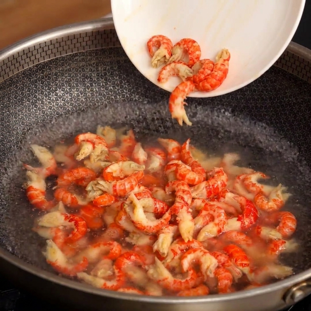 Un plato blanco inclinándose para verter las colas de cangrejo peladas en una sartén con agua hirviendo.