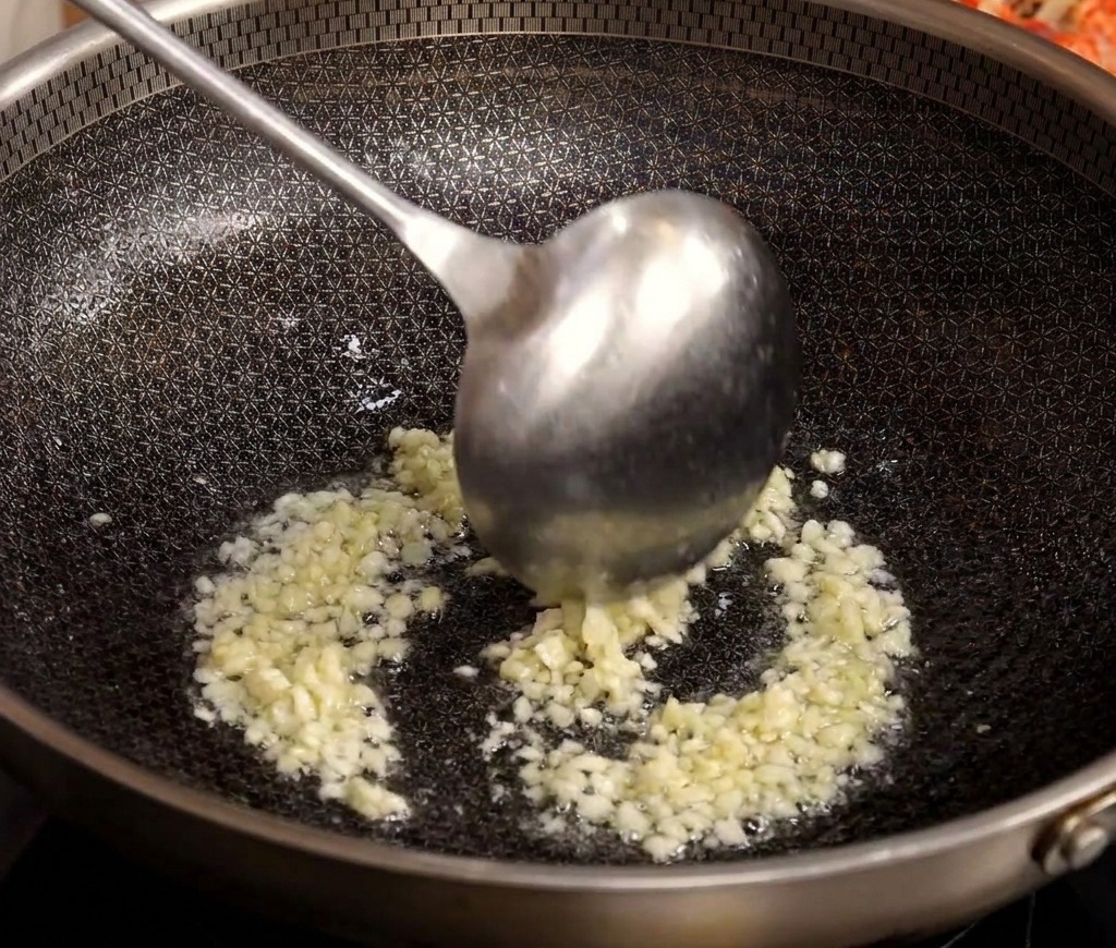Un cucharón metálico removiendo ajo picado mientras se dora en un wok caliente con aceite.