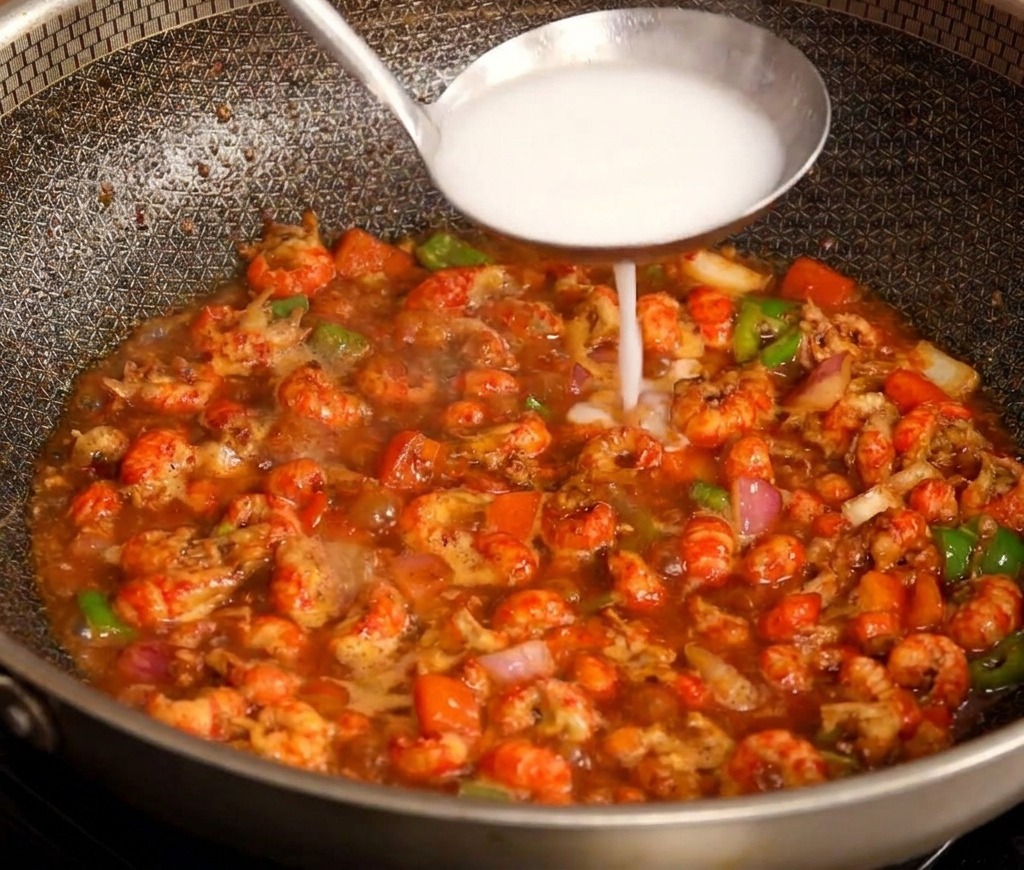 Un cucharón de acero inoxidable vertiendo una mezcla blanca de almidón en el centro de un salteado de cangrejo rojo.