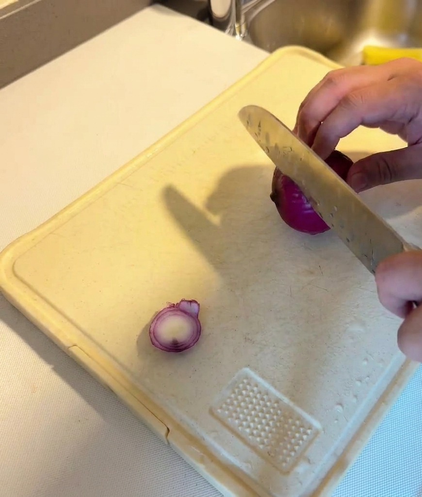 Una persona cortando una cebolla roja en láminas finas sobre una tabla de cortar blanca.