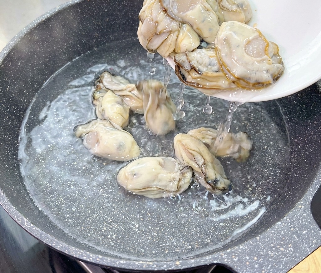 Ostras frescas y crudas se deslizan desde un plato blanco hacia un wok con agua fría.