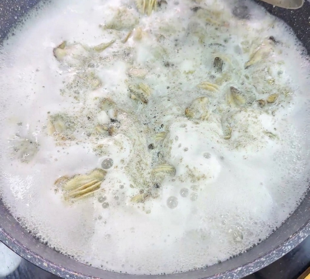 Un colador metálico levanta ostras escalfadas de una olla con agua hirviendo vigorosamente y con espuma.