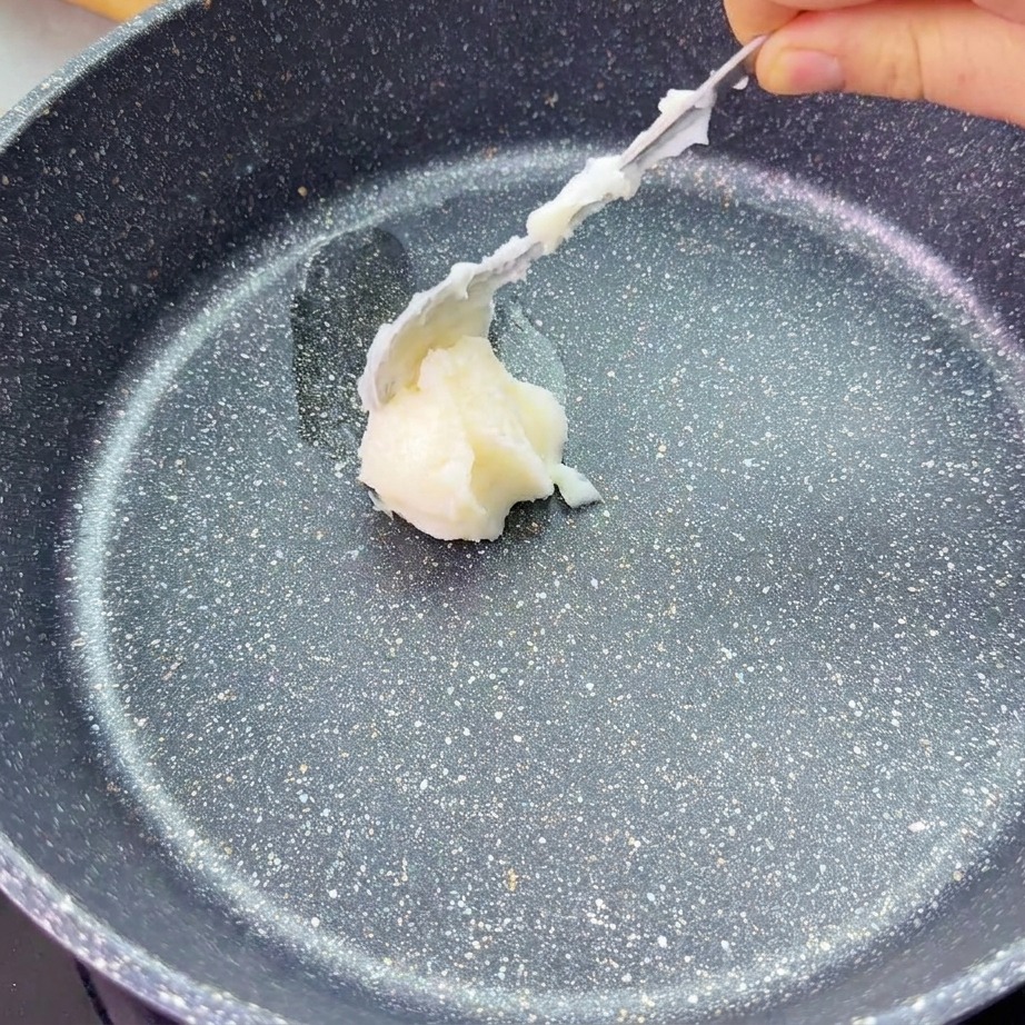Una cuchara coloca una porción de manteca blanca en el centro de una sartén oscura con acabado de piedra.