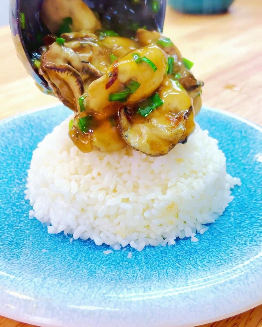 Un cucharón sirve la salsa espesa y las ostras sobre un montículo de arroz blanco en un plato de cerámica azul.