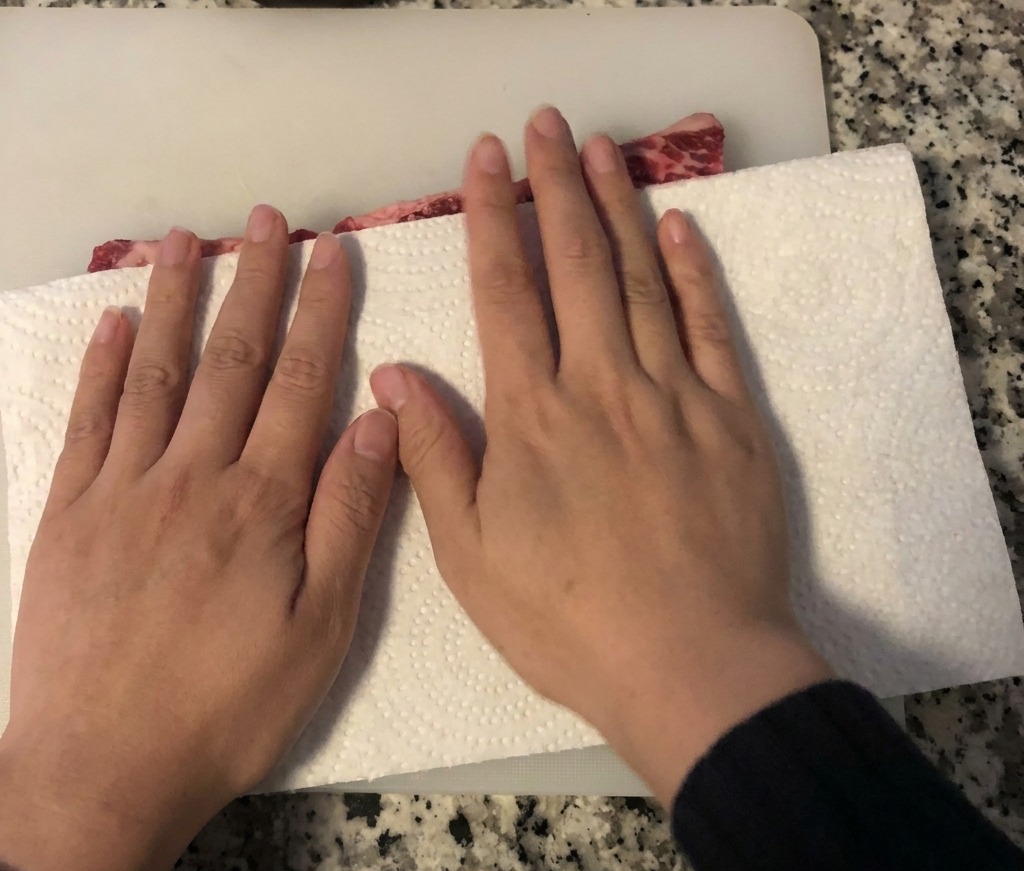Manos usando papel absorbente para secar las costillas de res crudas sobre una tabla de cortar blanca.