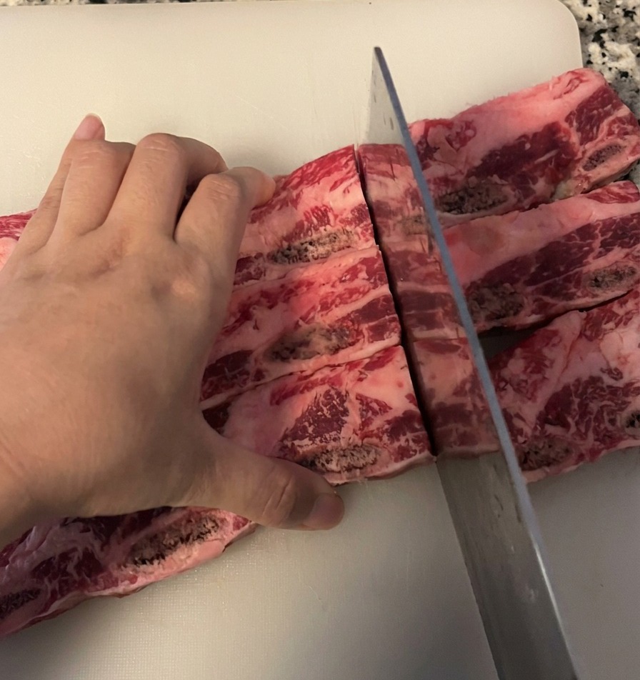 Una persona cortando un trozo de costillas de res en porciones individuales con un cuchillo de chef sobre una tabla blanca.