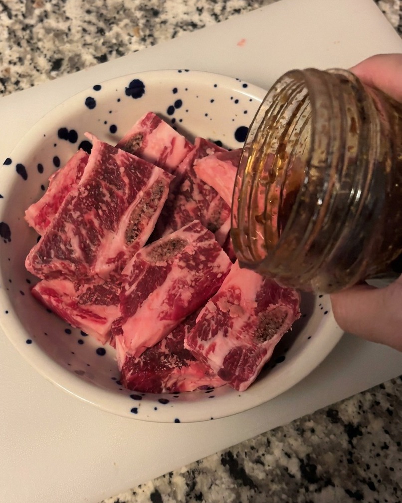 Salsa oscura y brillante para barbacoa coreana vertiéndose desde un frasco sobre costillas de res crudas en un bol blanco.
