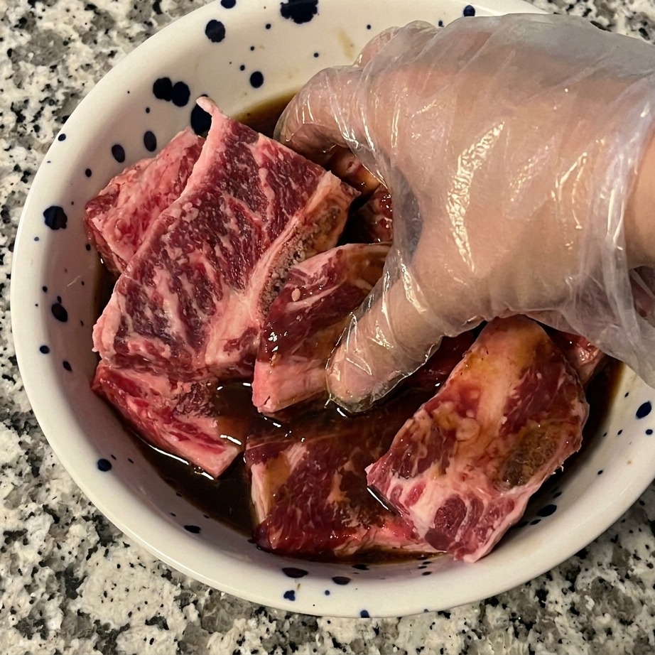 Una mano con un guante de plástico transparente mezclando las costillas de res con una marinada oscura en un bol.