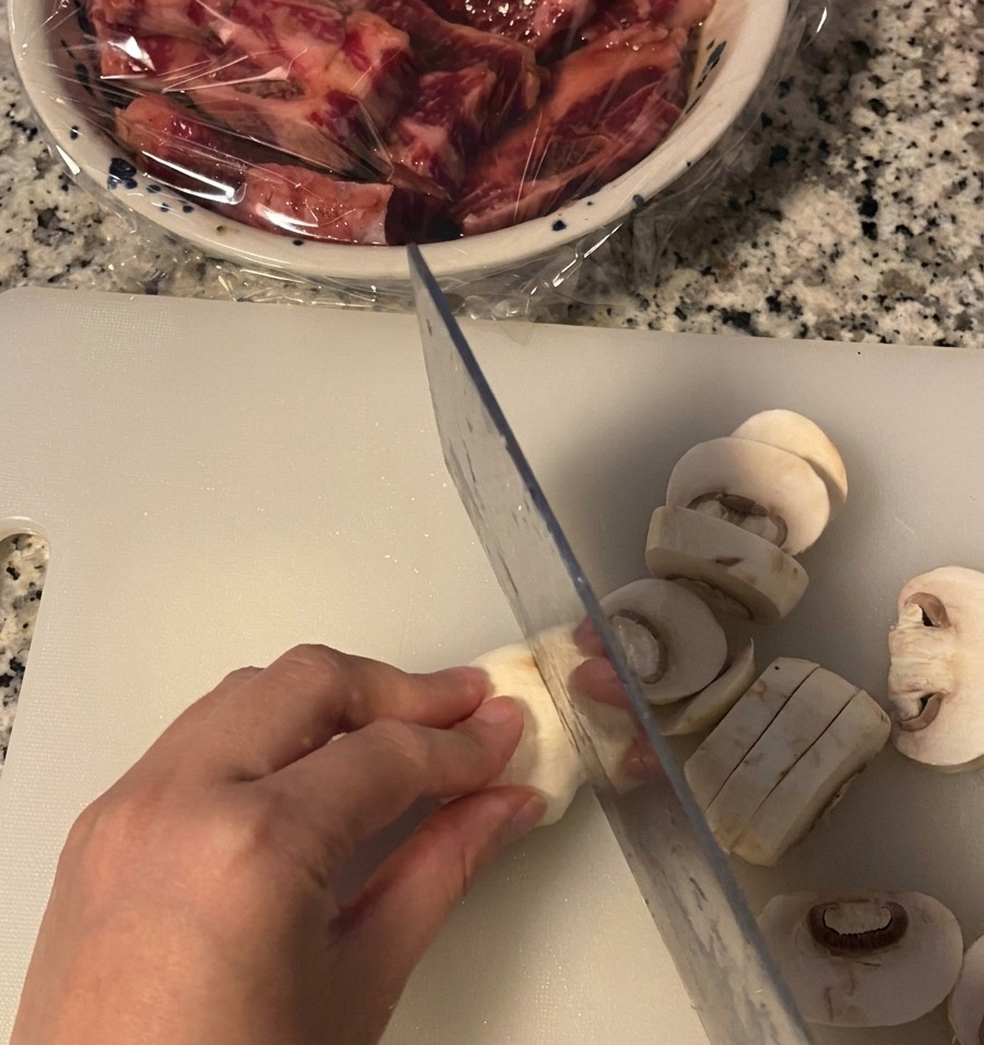Una mano sujetando un champiñón blanco mientras lo corta en rodajas gruesas con un cuchillo sobre una tabla de cortar.